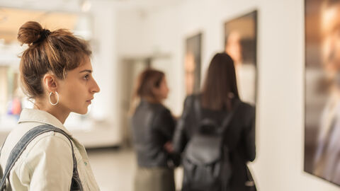 Symbolbild Museum Am linken Bildrand ist eine Frau mit einem Dutt, in einer hellen Jacke und mit einer Tasche über der Schulter zu sehen, die sich ein Gemälde ansieht, dass am rechten Bildrand angeschnitten ist. Im Hintergrund sind zwei weitere Frauen in dunklen Jacken zu sehen, die sich andere Gemälde ansehen.