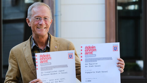 Staatsminister Wintermeyer hält zwei Förderbescheide "#deinehrenamt" nebeneinander in die Kamera und lächelt.