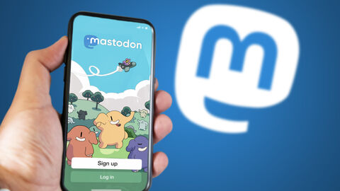 Vor einem blauen Hintergrund hält eine Person ein Smartphone in der Hand. Auf dem Bildschirm ist die App Mastodon zu sehen.