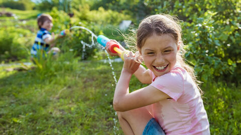 Zwei lachende Kinder bespritzen sich auf eienr Wiese mit Wasser.