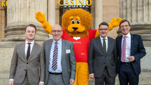Vier Männer stehen stehen vor einem Gebäude. In der Mitte steht eine Person als plüschiger Löwe angezogen.