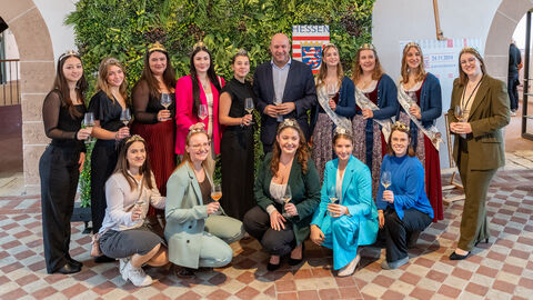 mehrere junge Frauen stehen gemeinsam mit einem Herren vor einer Wand mit Pflanzen und schauen in die Kamera. Alle halten ein Weinglas in den Händen.