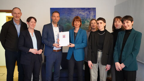 Sozialministerin Hofmann steht mit weiteren Personen zum Gruppenfoto aufgestellt in einem Raum. Sie übergibt einen Förderbescheid.