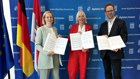 Zwei Frauen und ein Mann stehen vor einer blauen Wand und halten Dokumente hoch.