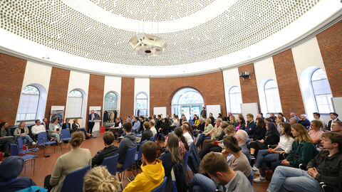 Großer, runder Saal mit Menschen, die auf Stühlen sitzen.