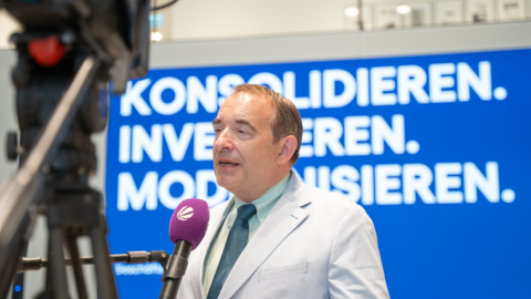 Herr Lorz spricht in ein Mikrofon mit dem Symbol von Sat1 - vor ihm sind Kameras aufgebaut, im Hintergrund die Worte "Konsolidieren, Investieren, Modernisieren"