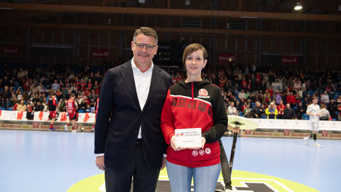 Ministerpräsident Boris Rhein steht auf dem Spielfeld der MT Melsungen. Die Ehrenamtliche Jessica Fetzer hat von ihm eine Auszeichnung überreicht bekommen. Diese hält sie in der Hand.