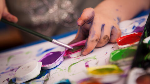 Hand eines Kindes mit Wasserfarbe verschmiert und Pinsel in der Hand an einem Farbkasten