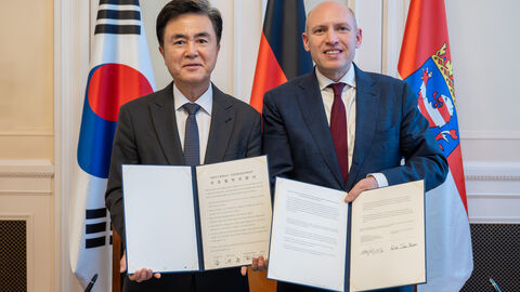 2 Männer stehen nebeneinander und halten jeweils ein Dokument in die Kamera.