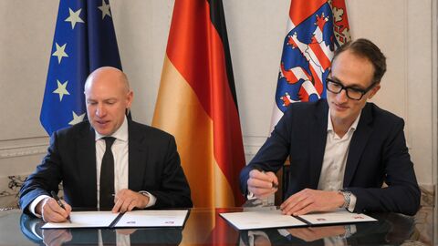Zwei Personen sitzen an einem Tisch und unterzeichnen Unterlagen. Im Hintergrund sind die Flaggen von Europa, Deutschland und Hessen aufgestellt.