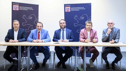 Fünf Personen sitzen an einem Tisch, im Hintergrund Plakate mit dem Wappen des Hessischen Ministeriums für Wissenschaft und Forschung, Kunst und Kultur