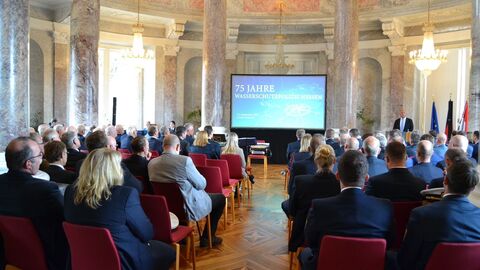 Stuhlreihen mit Menschen schauen auf eine Wand, wo eine Präsentation drauf projiziert wird.