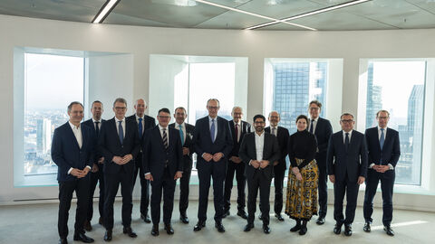 Eine Gruppe von Menschen stehen in einem Raum in einem Hochhaus. Im Hintergrund ist ein anderes Hochhaus aus dem Fenster zu sehen.